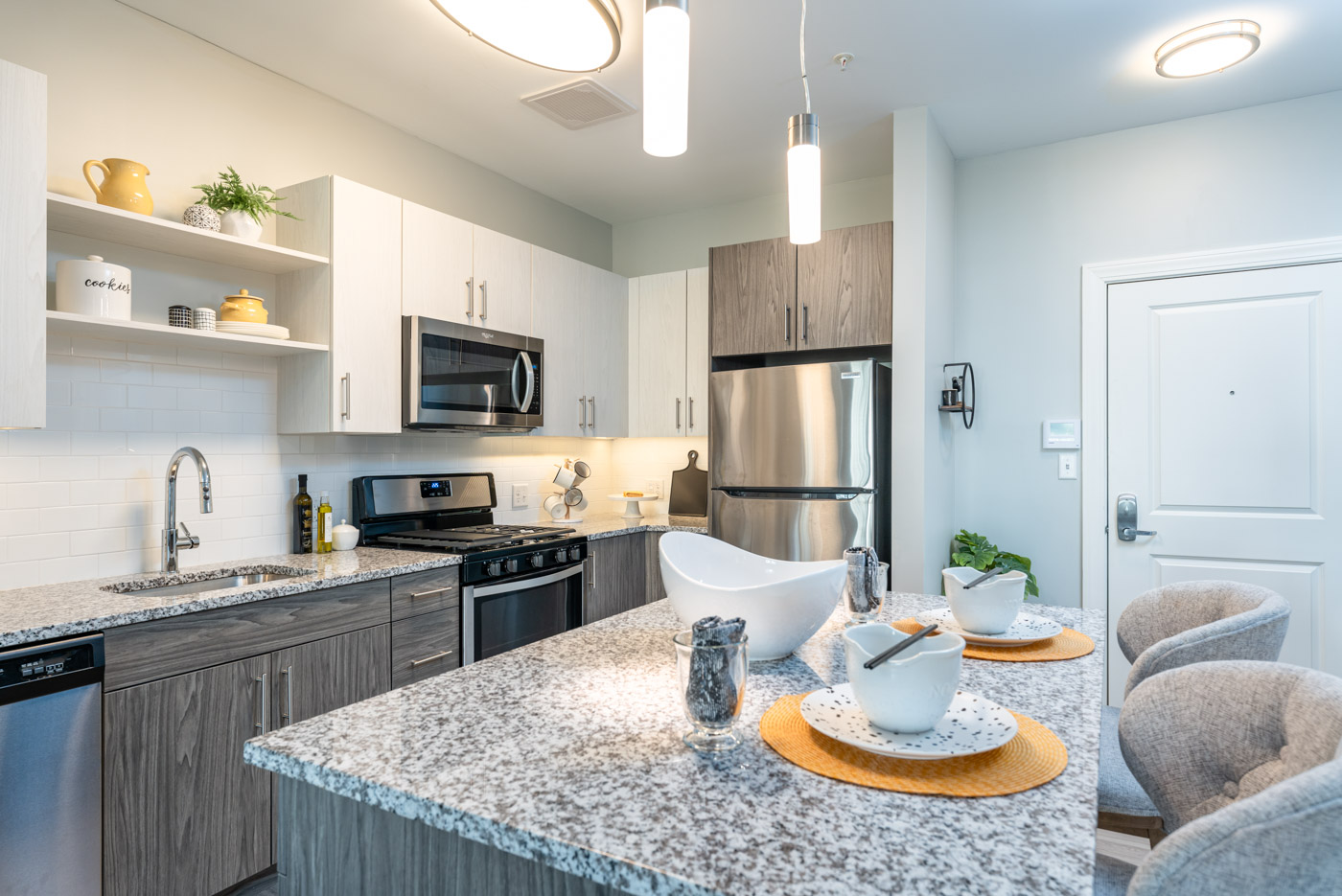 One bedroom model apartment kitchen with stainless appliances, marble counters, white cabinets, stove, microwave and oven with undercabinet lighting. Kitchen island with place settings and overhead lights